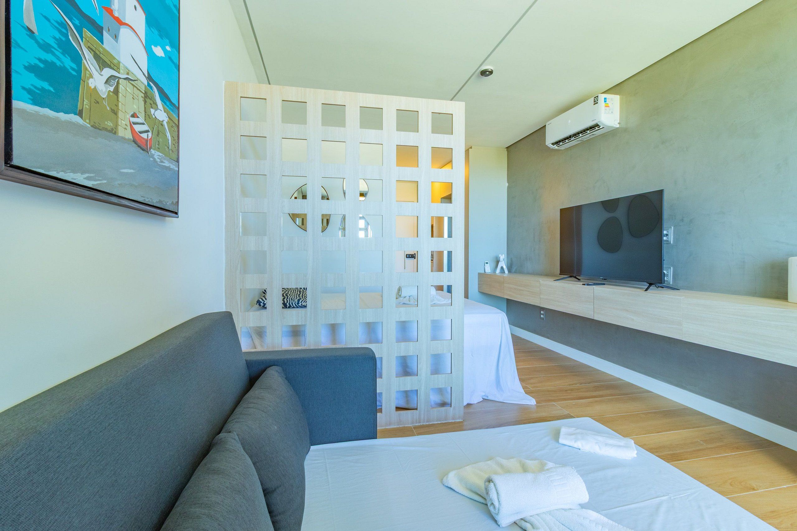 Quarto de hotel moderno com sofá, cama com toalhas, televisão e piso de madeira.