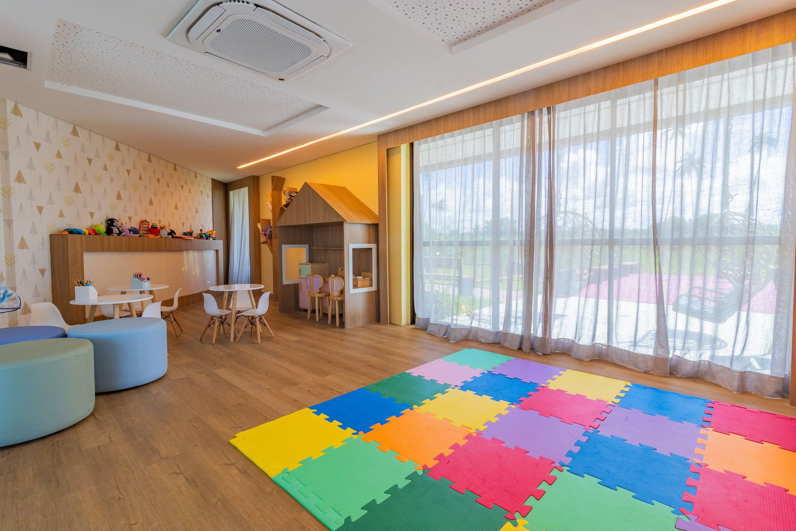 Una habitación moderna con piso de madera, alfombras de espuma coloridas y un techo blanco, y un ventanal con cortinas translúcidas.