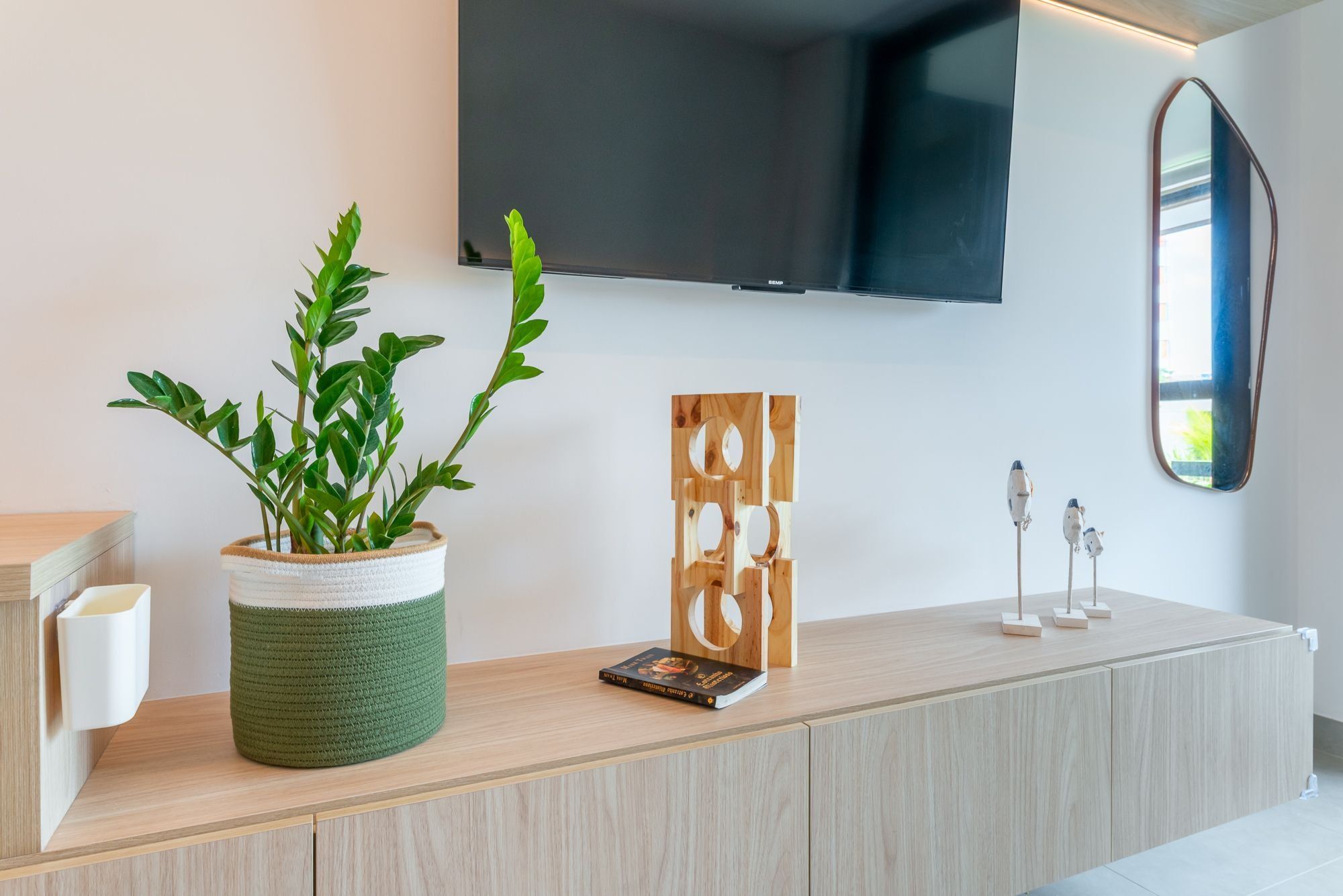 A modern living room with a wooden cabinet, a green plant in a woven basket, a framed artwork, and a mounted television.
