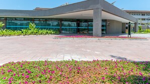 A modern building with a curved roof, large glass windows, and a paved entrance area with flower beds. - Beach Class Summer by Carpediem (Ipojuca)