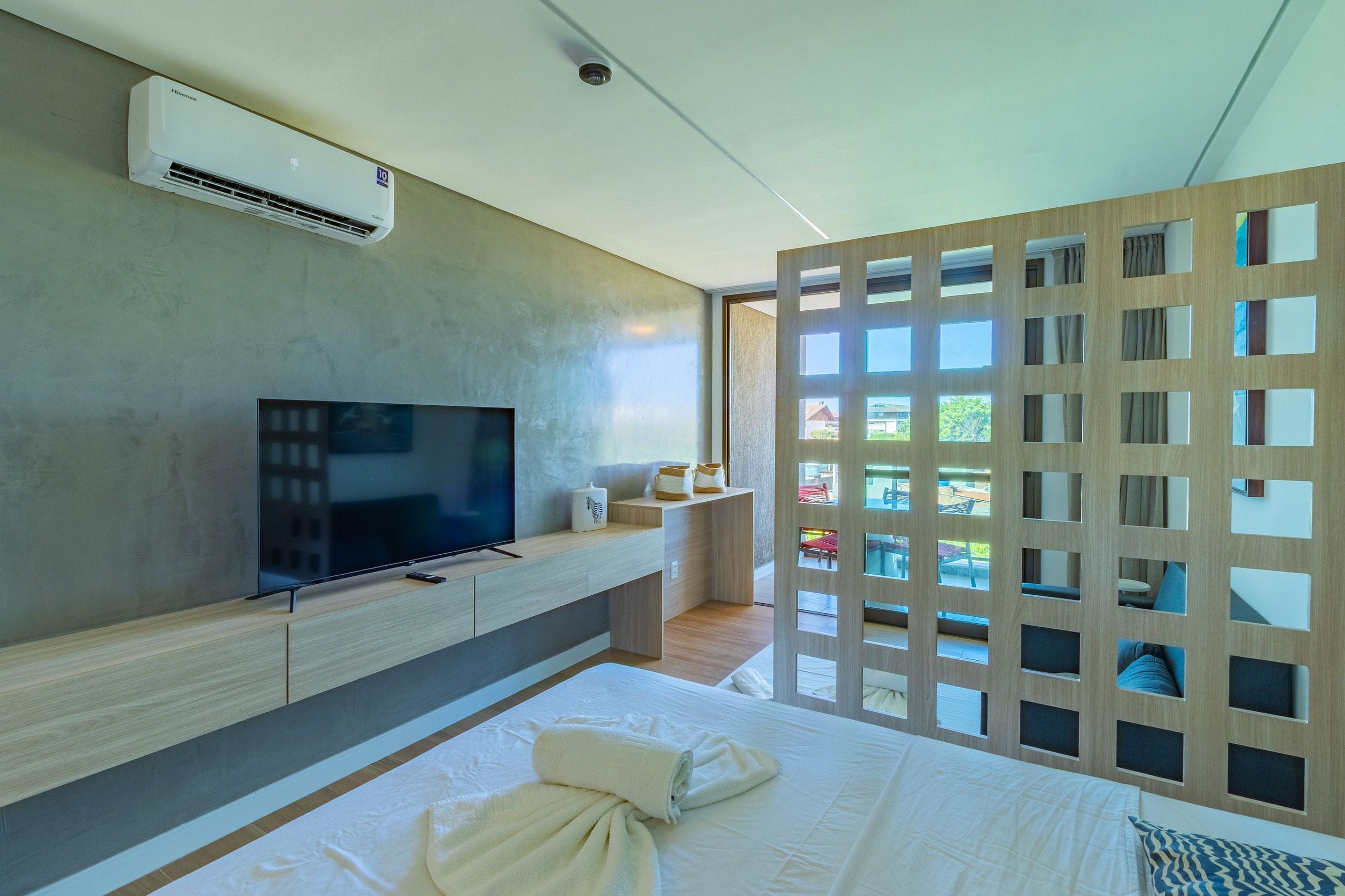 Quarto de hotel moderno com televisão de tela plana, divisória de madeira com várias janelas, cama com toalhas dobradas e ar-condicionado no teto.