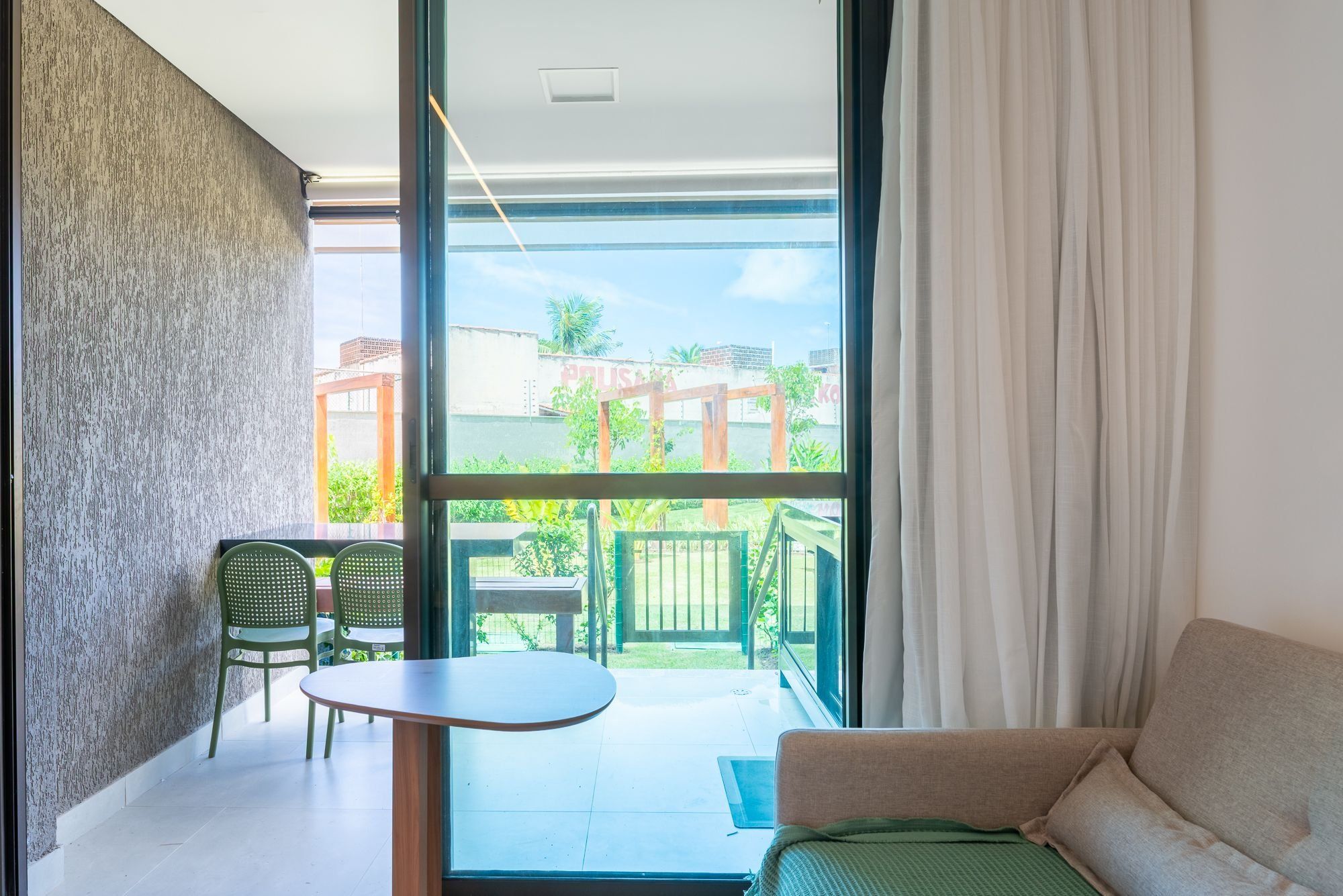 Een moderne woonkamer met een bank, een kleine tafel en twee stoelen. Een groot raam biedt uitzicht op een balkon met groen en gebouwen.