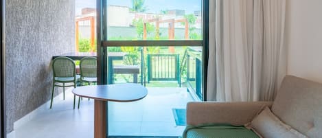 A modern living room with a sofa, a small table, and two chairs. A large window offers a view of a balcony with greenery and buildings.