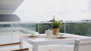 Een witte tafel met een potplant, theepot, kopjes en een placemat op een balkon met glazen hekwerk en uitzicht op groen.