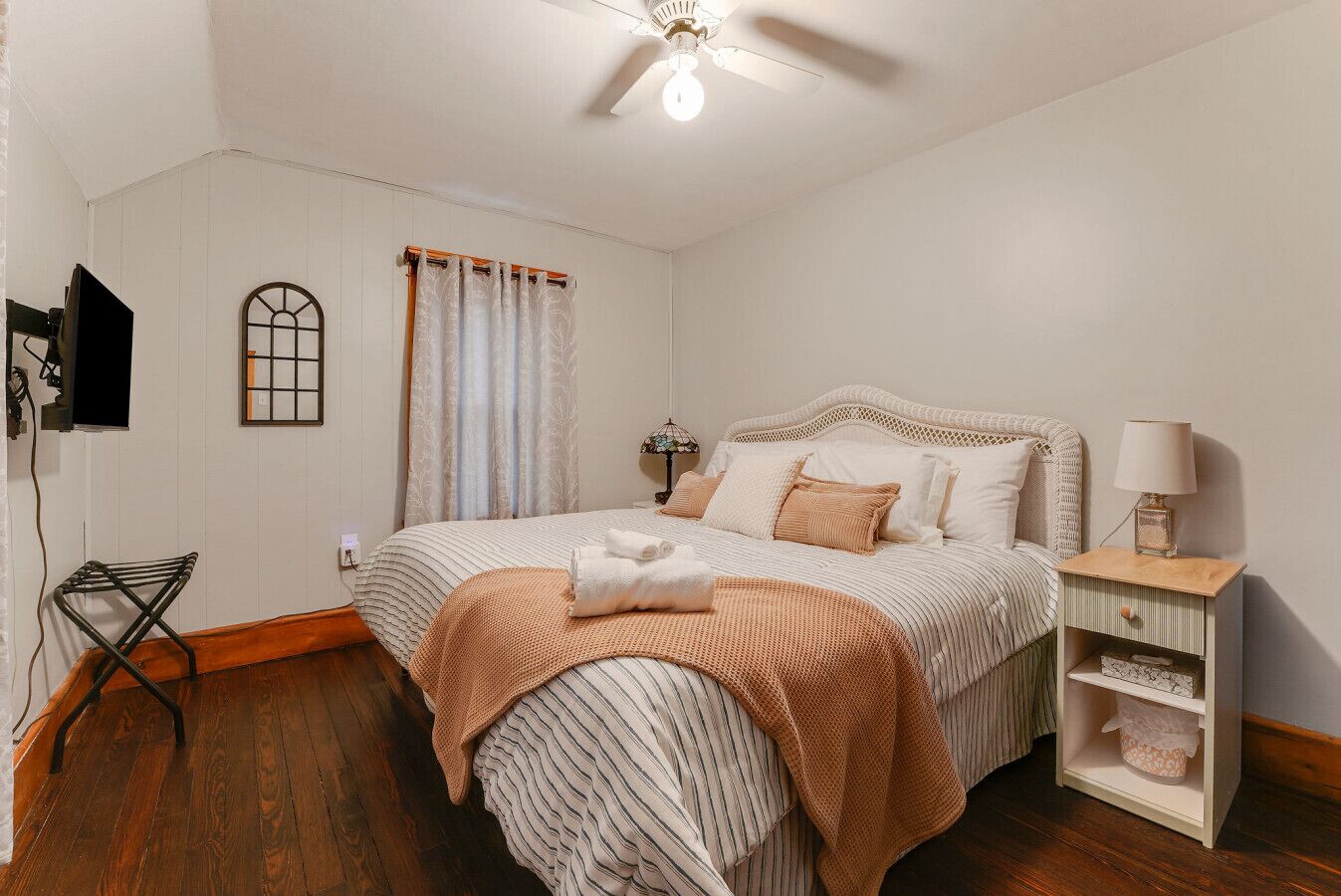 Une chambre à coucher comprenant un lit, une table de chevet, une lampe, une fenêtre avec des rideaux, un téléviseur fixé au mur et un ventilateur de plafond.