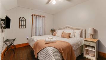 Une chambre à coucher comprenant un lit, une table de chevet, une lampe, une fenêtre avec des rideaux, un téléviseur fixé au mur et un ventilateur de plafond.