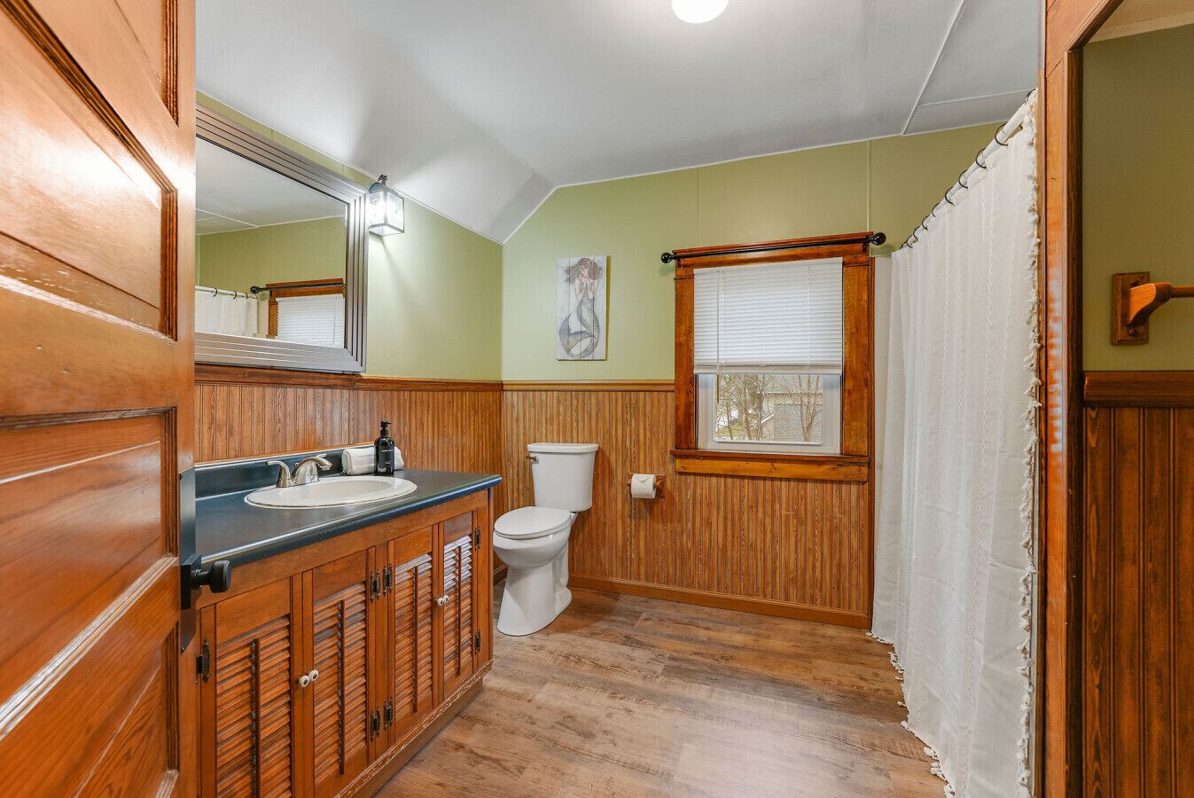 Une salle de bain avec des lambris en bois, un lavabo, des toilettes et une fenêtre avec des stores.