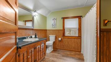 Une salle de bain avec des lambris en bois, un lavabo, des toilettes et une fenêtre avec des stores.