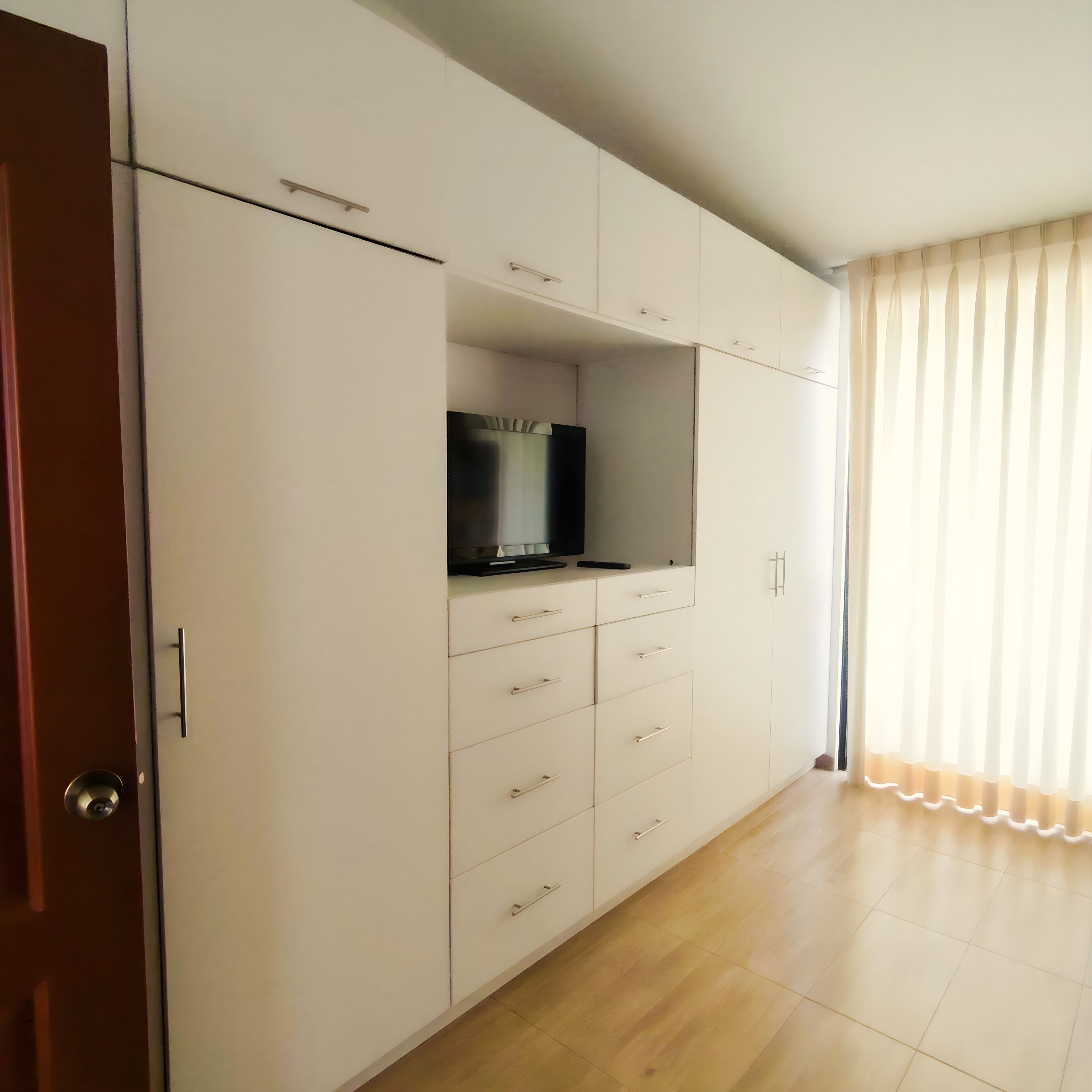 A room with a wardrobe, a television on top, and a large window with curtains.