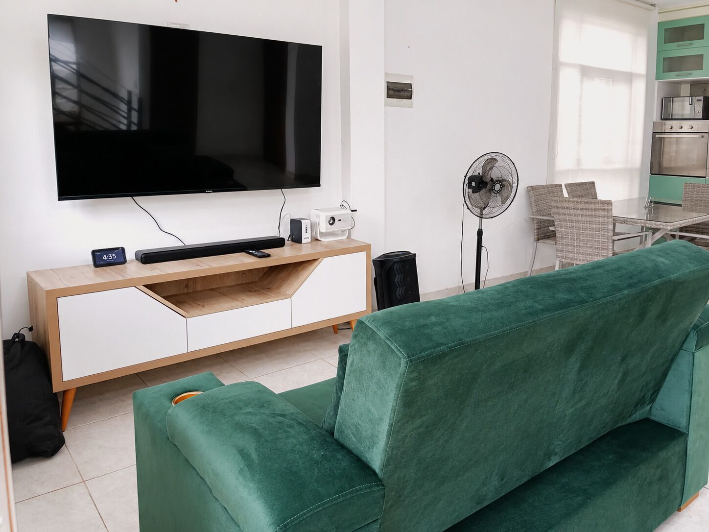 A living room with a flat-screen TV, a wooden entertainment unit, a green sofa, a standing fan, and a kitchen area with a microwave and oven.