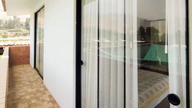 Una habitación con puerta corrediza de vidrio que da a un balcón, una cama con colcha estampada y vista a un paisaje con árboles y colinas.