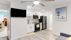 Uma cozinha moderna com armários brancos, uma geladeira preta e um micro-ondas. Há um ventilador de teto e um quadro com uma cena de praia na parede.