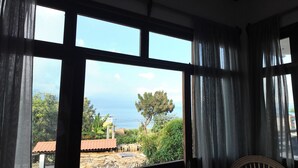 Una habitación con ventanas amplias que ofrece vistas a un paisaje costero, incluyendo árboles y un edificio con techo rojo.