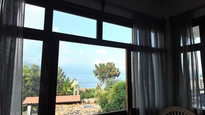 Una habitación con ventanas amplias que ofrece vistas a un paisaje costero, incluyendo árboles y un edificio con techo rojo.