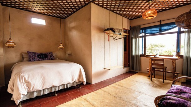 Ein Schlafzimmer mit einem Bett, einem Schreibtisch und einem Stuhl. Es gibt ein Fenster mit Vorhängen, eine Hängeleuchte und einen Teppich auf dem Boden.