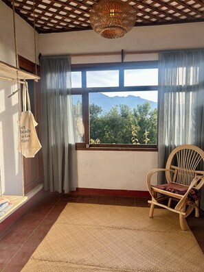 Una habitación con un gran ventanal, una silla de mimbre, un bolso colgado y una alfombra tejida.