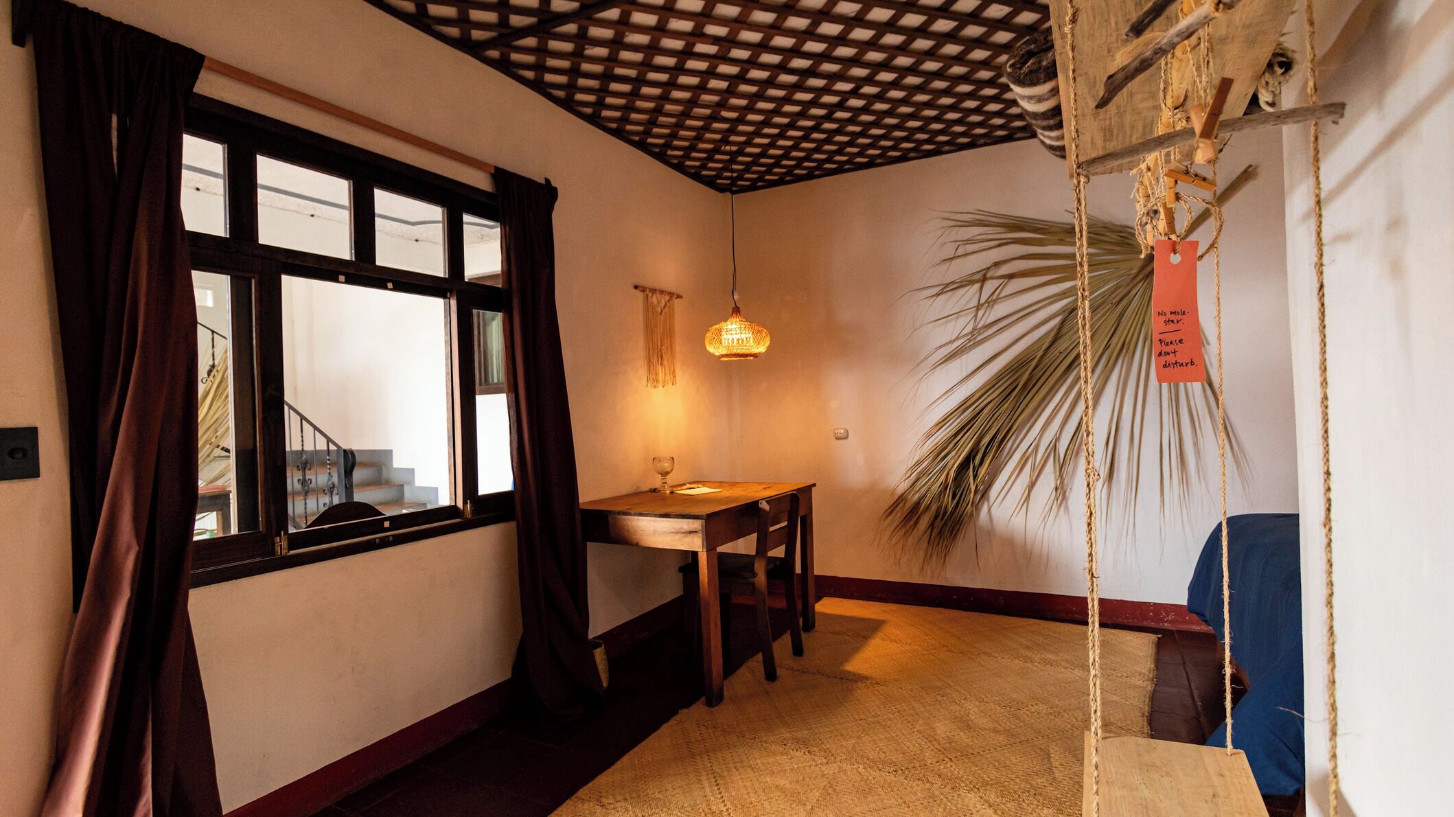 A room with a wooden table, a hanging light fixture, a large window with curtains, and a palm plant.