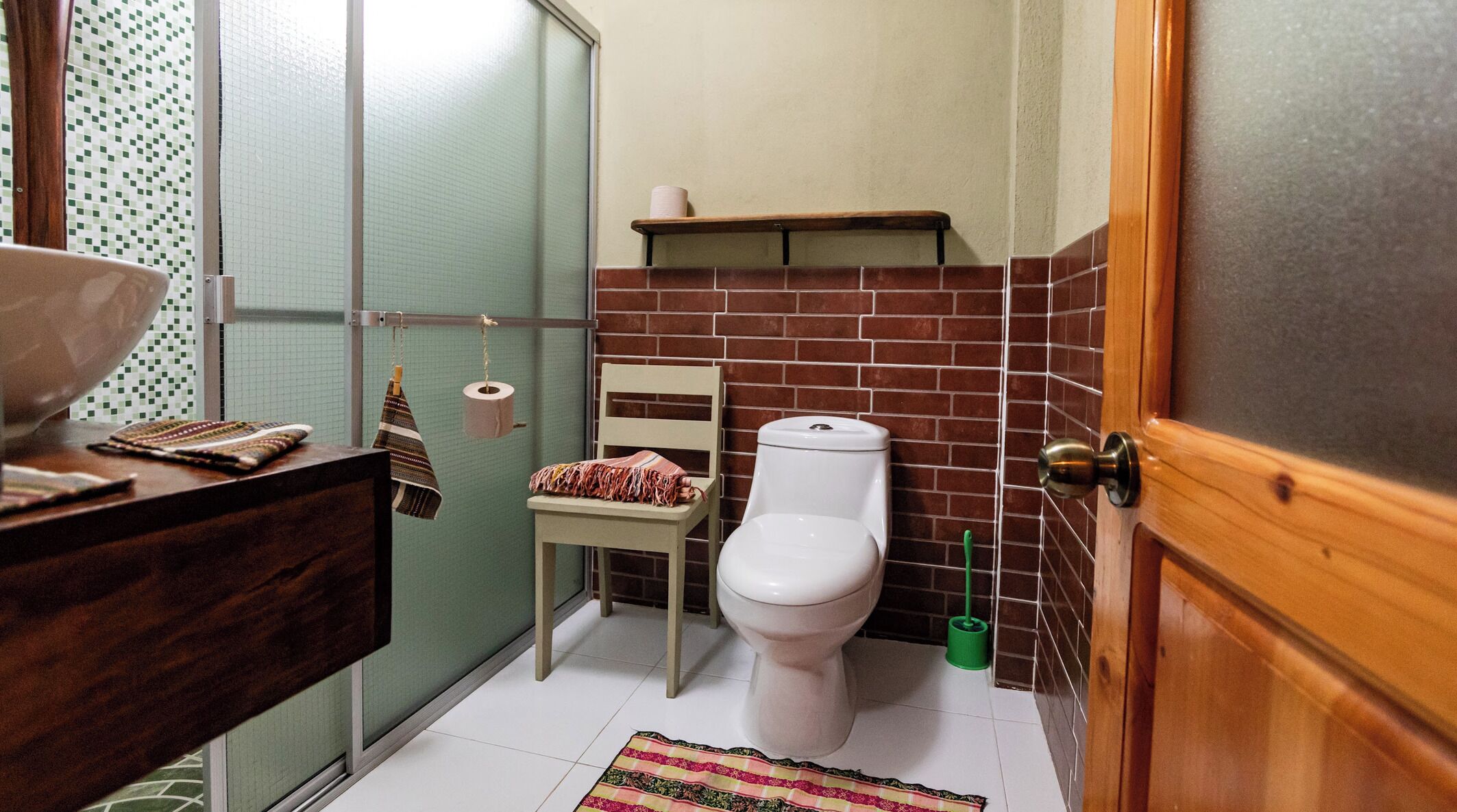 A compact bathroom with a toilet, a small chair, a shelf above the toilet, and a shower area.