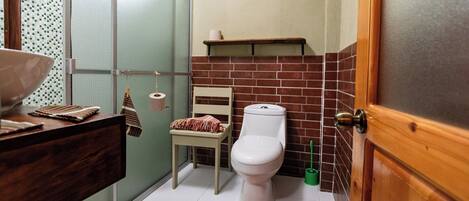 A compact bathroom with a toilet, a small chair, a shelf above the toilet, and a shower area.