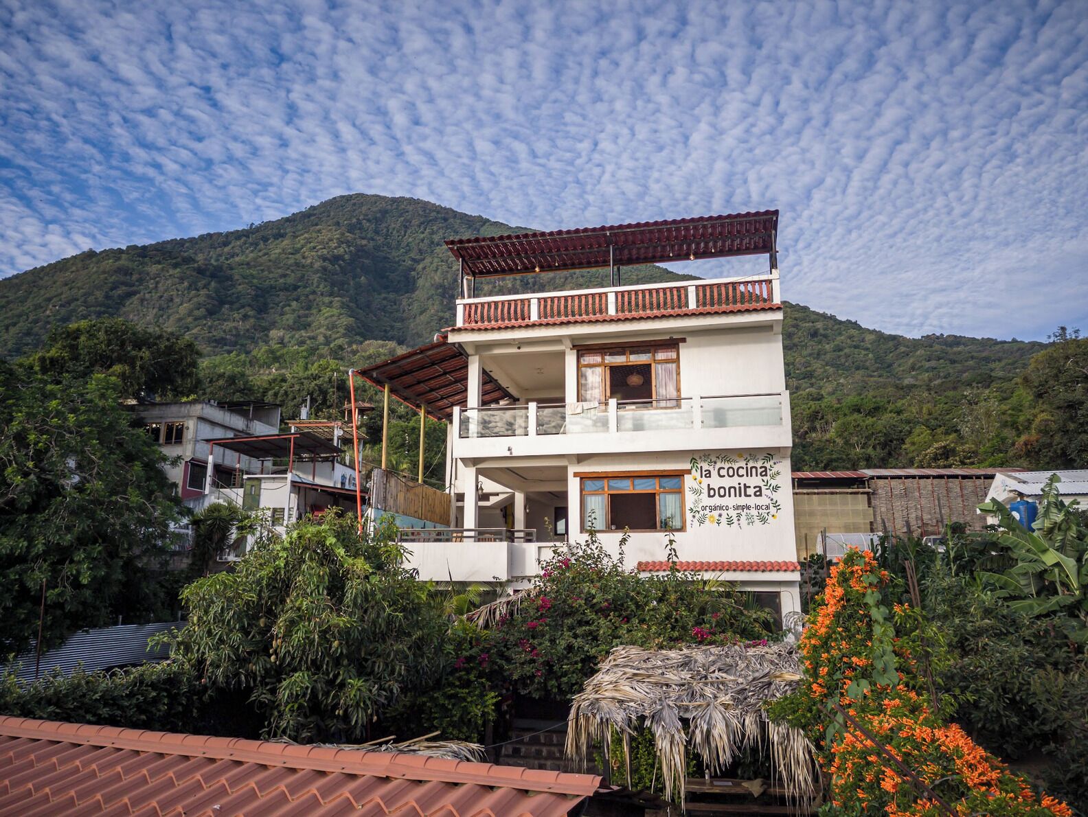 Un bâtiment à deux étages doté d’un balcon, entouré d’une végétation luxuriante et avec une montagne en arrière-plan.