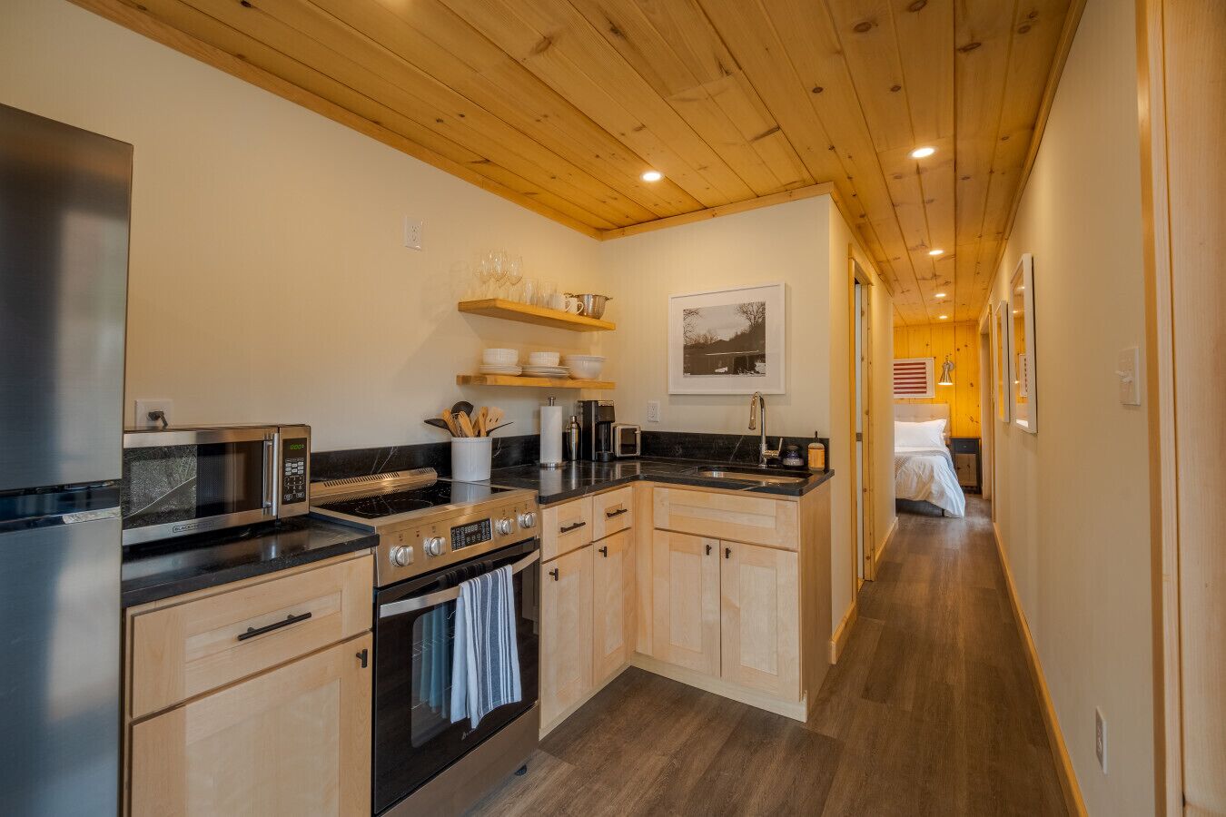 Cocina moderna con armarios de madera, un refrigerador de acero inoxidable, microondas y estufa. Hay una repisa de madera con vajilla y un cuadro en la pared.