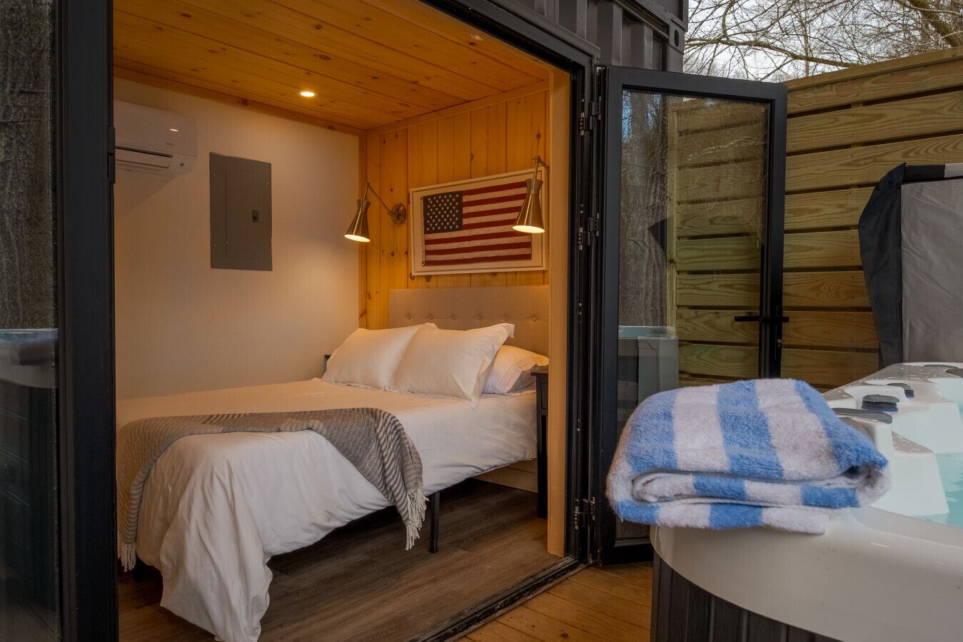 Une chambre confortable avec un lit, un drapeau américain accroché au mur et une fenêtre donnant sur une terrasse en bois.