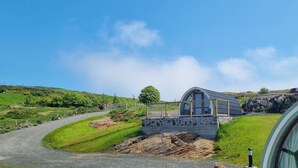 A modern house with a stone base and a glass dome, surrounded by a lush green landscape and a gravel path.