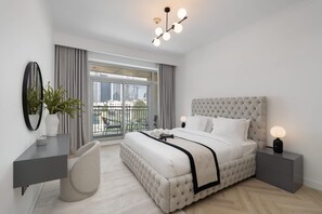 A modern bedroom with a tufted headboard, a large bed, a desk, a chair, a bedside table with a lamp, and a view of the city through large windows. - Stayshort - Luxury 1 Bedroom in Downtown With Burj Khalifa View (Dubai)