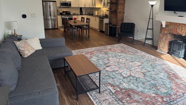 A modern living room with a gray sofa, a wooden coffee table, and a patterned rug. A kitchen with white cabinets and a brick accent wall is visible in the background.