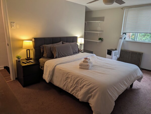A neatly made bed with a white comforter and pillows, a bedside table with a lamp, a small plant, and a radiator.