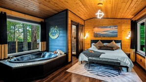 Ein Holzblockhaus-Innenraum mit einem großen Bett, einer Whirlwanne und einem Fenster mit Blick auf Bäume.