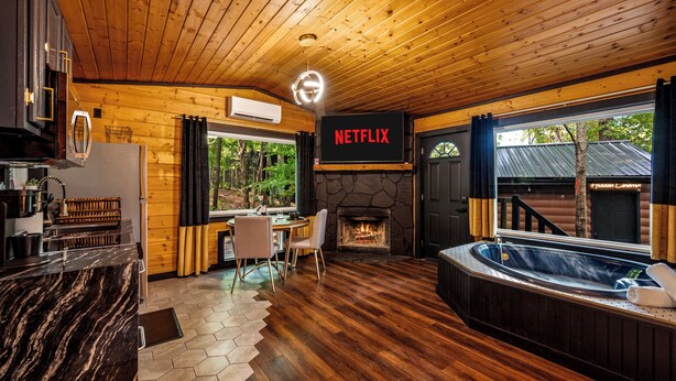 Ein gemütliches Blockhaus-Interieur mit Kamin aus Stein, Whirlpool, Netflix-Beschilderung, Holzdielen und einem Essbereich.