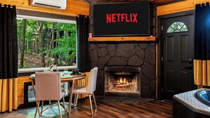 Ein gemütliches Zimmer mit Kamin aus Stein, einem Netflix-Schild, einem Essbereich mit Tisch und Stühlen und Blick auf den Wald durch ein Fenster.