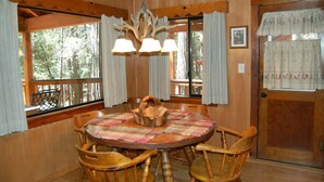 A wooden dining room with a round table, chairs, a chandelier, and large windows with curtains. - Deer Den (Yosemite National Park)