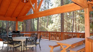 A covered deck with wooden furniture, a grill, and a view of tall trees. - Deer Den (Yosemite National Park)