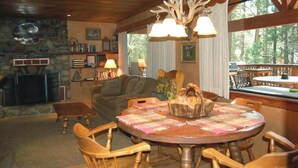 A cozy living room with a fireplace, wooden furniture, and a large window overlooking a forest. - Deer Den (Yosemite National Park)