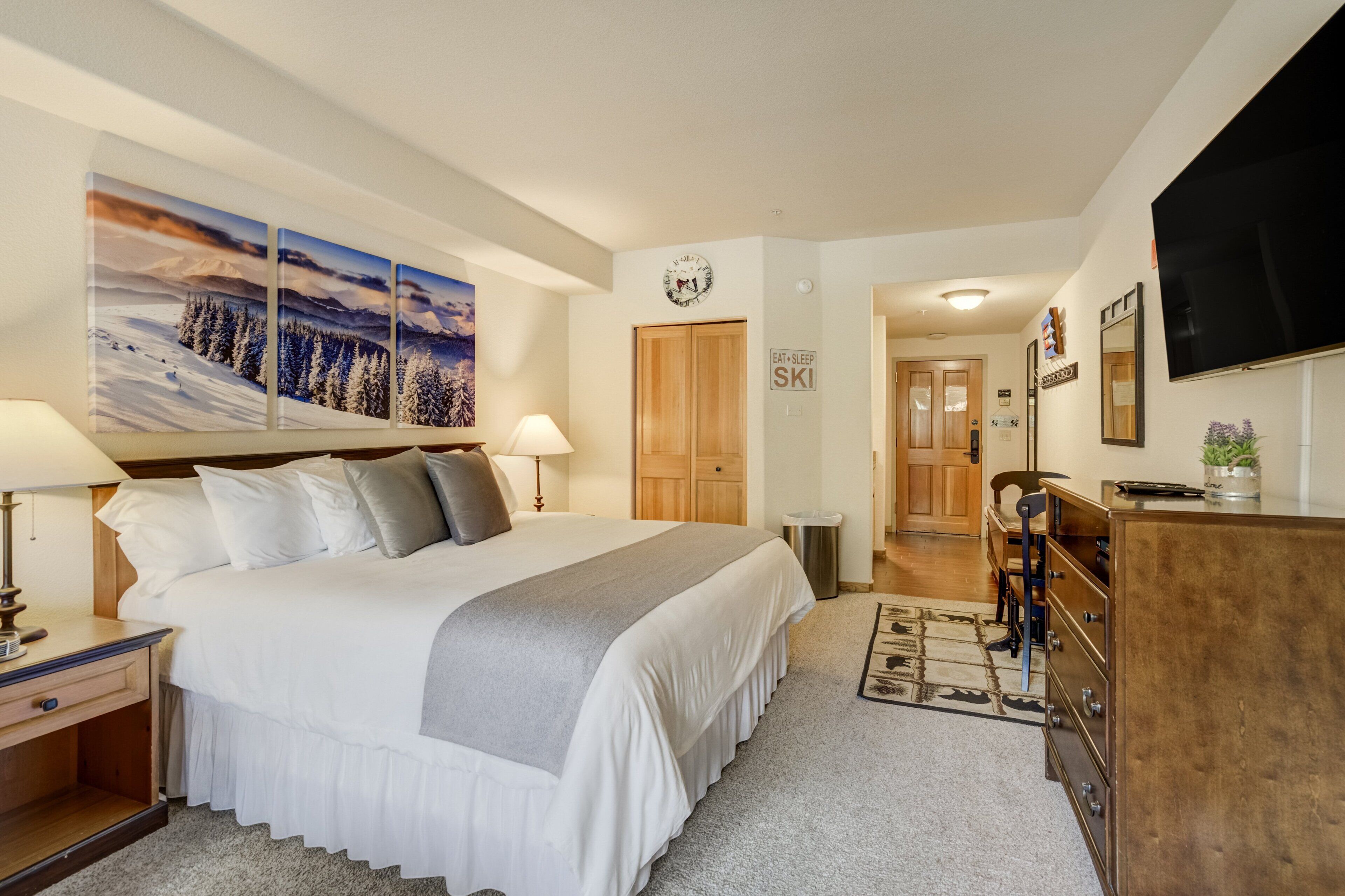 A hotel room with a large bed, a desk, a dresser, and a TV. There is a painting on the wall and a sign that says "Eat Sleep Ski".