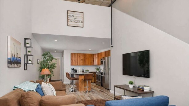Een moderne woonkamer met een bruine bank, een blauwe bank en een salontafel. Er is een keuken met houten kasten en een grote televisie aan de muur bevestigd.