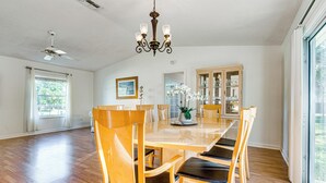 Un vaste espace dédié à la salle à manger, avec une grande table et des chaises en bois, un lustre et une vue sur l’extérieur grâce à de grandes fenêtres.