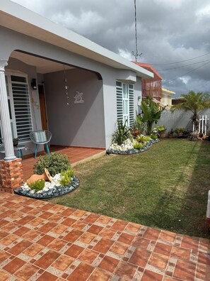 A modern house with a covered patio, a tiled patio, and a well-maintained garden with various plants and a small water feature. - Hernández Rental -complete house- (Aguadilla)