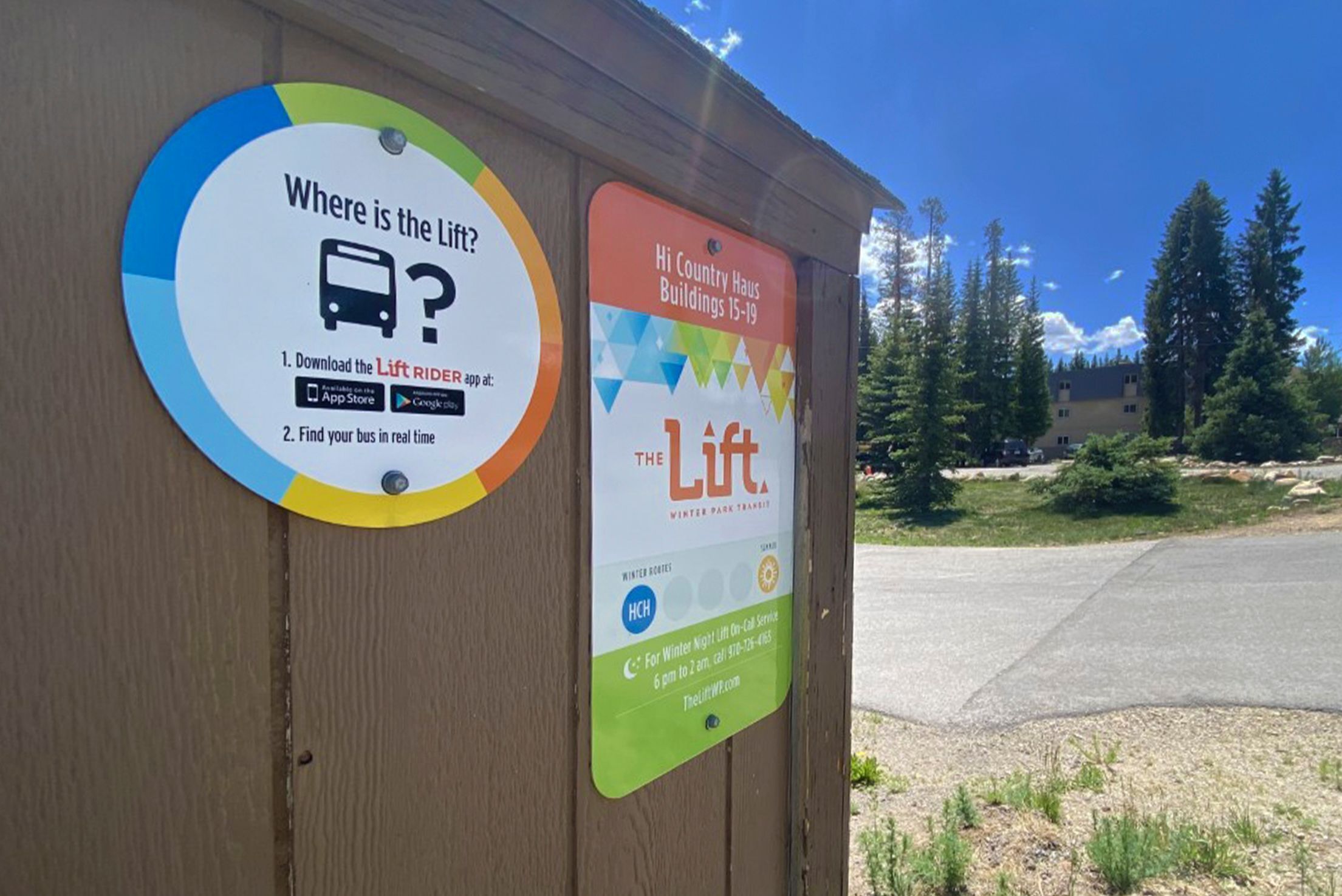 Un bâtiment en bois avec deux panneaux. L’un indique « Où se trouve l’ascenseur ? » et donne des instructions pour télécharger l’application Lift Rider. L’autre panneau indique « Hi Country Haus, bâtiments 15 à 19 » et mentionne un service de transport de Winter Park.