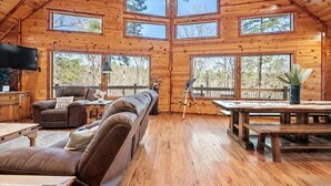 A spacious cabin with a high ceiling, multiple large windows, a cozy seating area with a sofa and armchairs, a dining table, and a mounted television. - Fantastic 5-bedroom/Hot Tub/Pool Table/Games/Fire Pit/Foosball/Shuffleboard (Broken Bow)