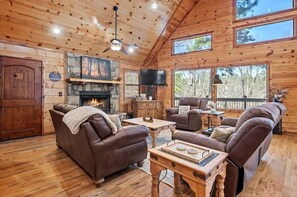 A cozy cabin interior with a fireplace, leather couches, wooden floors, and large windows. - Fantastic 5-bedroom/Hot Tub/Pool Table/Games/Fire Pit/Foosball/Shuffleboard (Broken Bow)