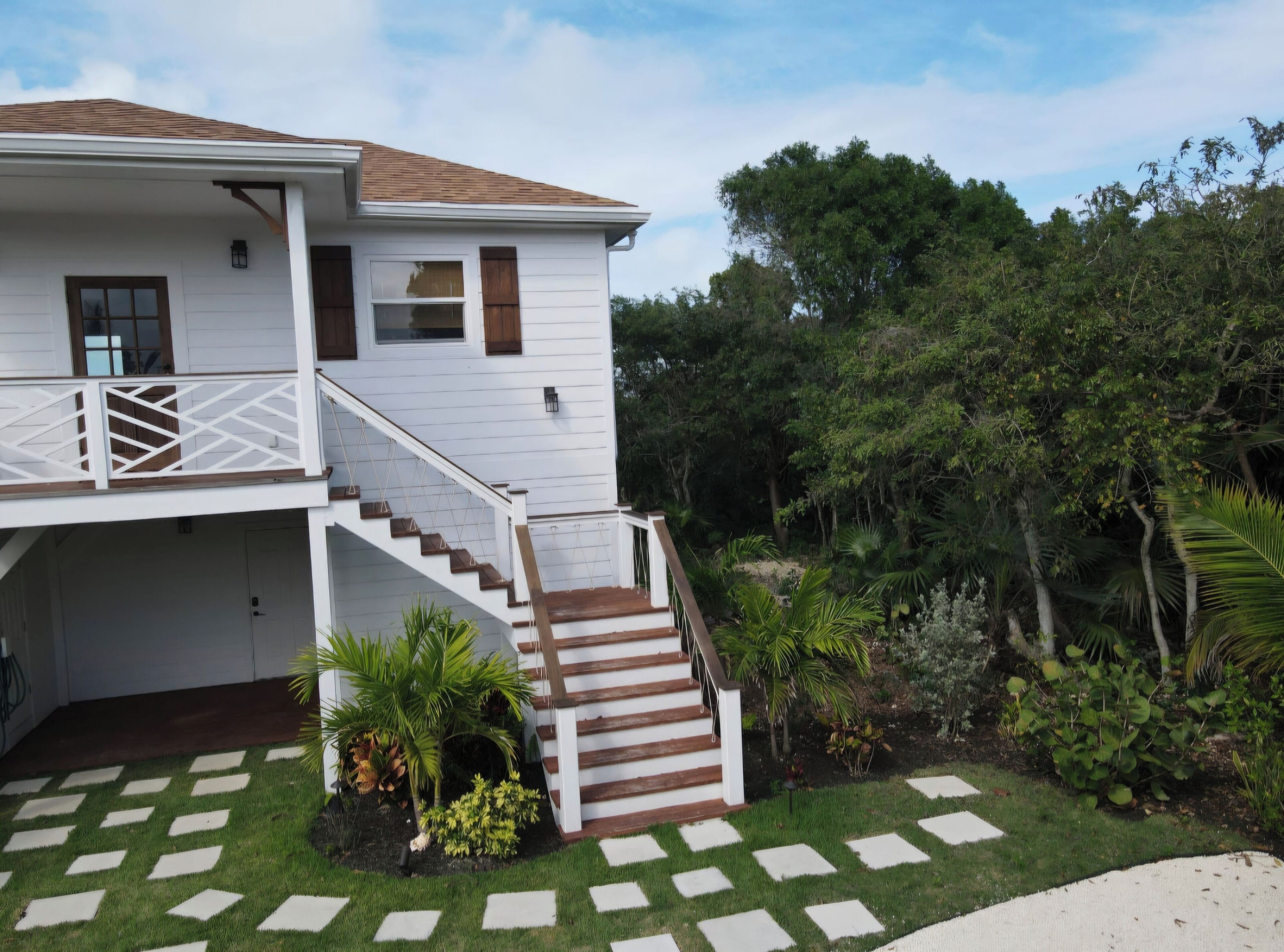 A two-story house with a wooden staircase, a balcony, and a garden with palm trees.