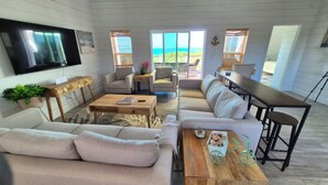 Ein geräumiger Wohnbereich mit Sofa, Couchtisch und Esstisch, einem an der Wand befestigten Flachbildfernseher und großen Fenstern mit Blick auf den Ozean.
