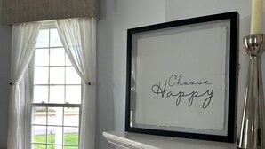 Una habitación con chimenea, un cartel enmarcado con el texto "Choose Happy", un sillón con cojín y una ventana con cortinas.