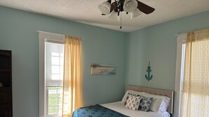 Un dormitorio con una cama, ventilador de techo, ventana con cortinas y un adorno fijado a la pared.