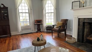 Un salón con pisos de madera, una chimenea, un sillón antiguo y una mesita con un letrero que dice 'hello'.