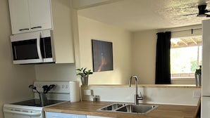 Eine kleine Küche mit weißen Schränken, einer Mikrowelle, einem Herd und einem Geschirrspüler. Ein Fenster mit Vorhang, ein Spülbecken und ein Topf mit Pflanze stehen auf der Arbeitsplatte.