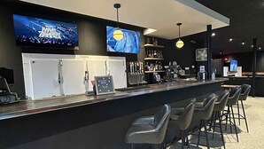 Zona de bar con una barra larga, varias televisiones mostrando contenido, una variedad de botellas en estantes y varios taburetes de bar.